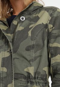 Maskovací bunda vyrobená z bavlněné směsi, s předním zipem, cvoky, elastickým pasem a zesílen�ými švy pro zvýšení odolnosti.