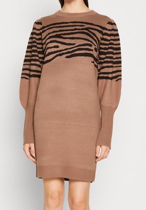 Kobieta ubrana w brązową, długorękawową dzianinową sukienkę za kolano z czarnymi pręgami zebry na klatce piersiowej i górnych partiach ramion.
