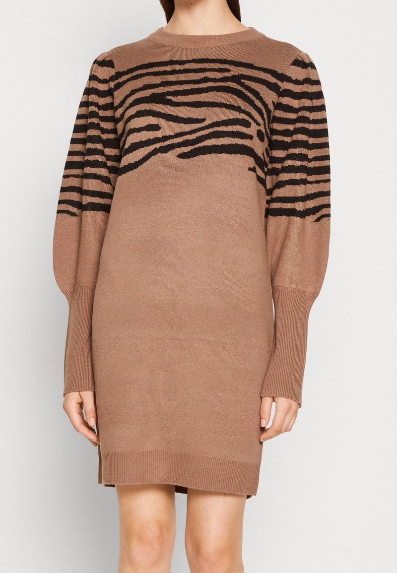 Femme portant une robe marron tricotée à manches longues et longueur genou, avec des rayures zébrées noires sur la poitrine et le haut des bras.