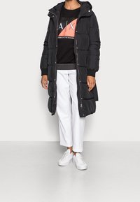 Svart puffjacka med huva, lång design, dragkedjestängning och kviltat mönster, kombinerad med en svart sweatshirt, vita korta byxor och vita sneakers.