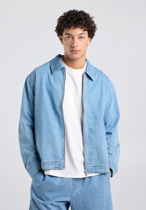 Jonge man met krullend haar, een lichtblauwe spijkerjas over een wit T-shirt en bijpassende lichtblauwe spijkerbroek, handen in de zakken.