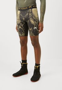 Shorts compressivi neri con una stampa dettagliata di teschi dorati e marroni, dotati di una vita con logo Venum. Indossati con scarpe alte nere e dorate.