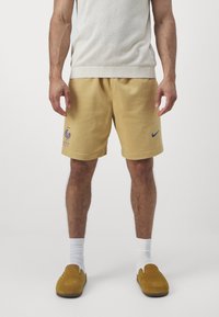 Mężczyzna ubrany w beżowe szorty sportowe z logo, białe skarpetki, musztardowe buty typu slip-on oraz koszulkę bez rękawów w jasnym beżu, stojący na tle jednolitego tła.
