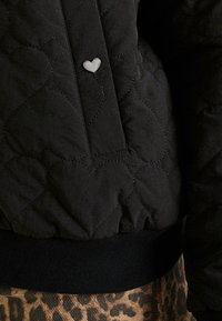 Chaqueta bomber de color negro acolchada con costuras en forma de corazón, un acento en forma de corazón plateado y una cintura de canalé negra.