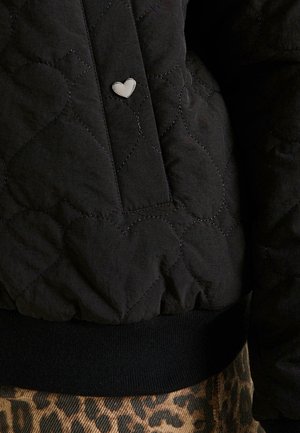 Veste bomber matelassée noire avec des coutures en forme de cœur, un accent en forme de cœur argenté et une taille en bord-côte noir.