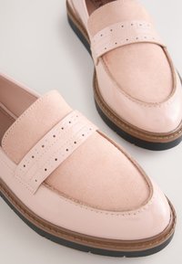 Paire de mocassins femmes rose clair avec textures en daim et cuir brillant, détail de bride perforée, et semelles en caoutchouc noir sur une surface blanche.