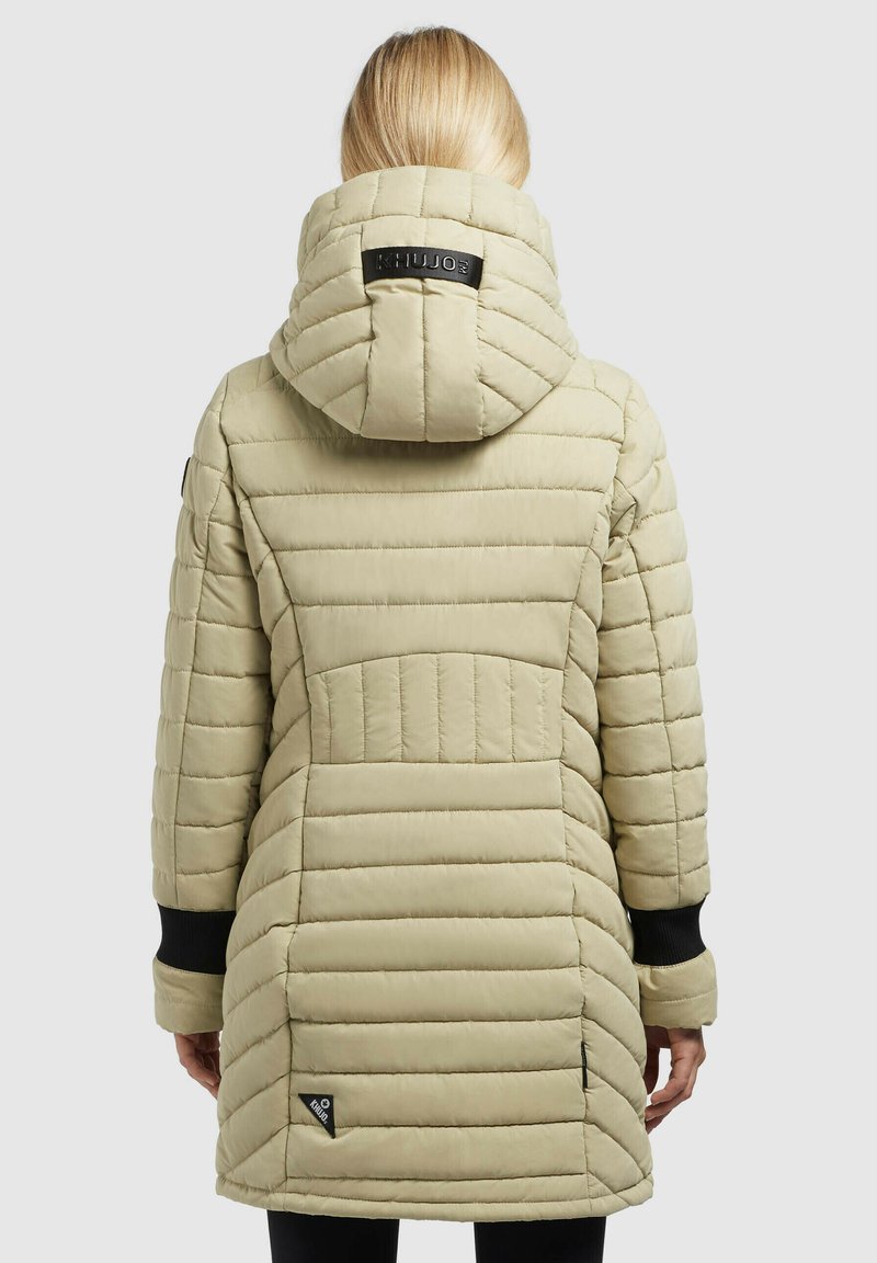 Frau, die eine beige gesteppte Parka mit Kapuze, schwarzen Handgelenkabschlüssen und Markenaufnähern an der oberen Kapuze und im unteren Rückenbereich trägt.