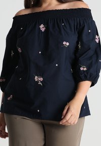 Femme portant un blouse bleu marine à épaules dénudées avec broderie florale rose et un pantalon beige, se tenant devant un fond neutre.