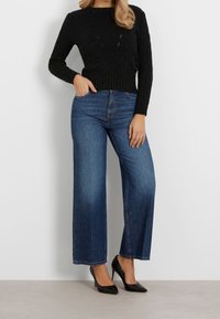 Suéter negro de punto cable con mezcla de lana, jeans de pierna ancha azul de talle alto y tacones negros de punta, contra una pared blanca.