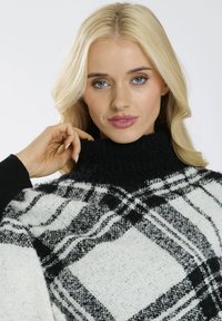 Pull à carreaux noir et blanc avec un col haut et pelucheux ; fabriqué en tissu doux, avec des manches ouvertes et une coupe décontractée.