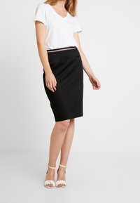 Svart pennkjol med slim fit och elastisk midja, kombinerad med en vit t-shirt med v-ringning och vita sandaler med remmar. Slät tygtextur.