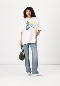 Camiseta blanca de gran tamaño con un gráfico colorido de cactus y un sol. Combinada con unos vaqueros acampanados en azul claro y zapatillas blancas.