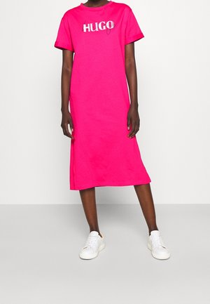 Persona che indossa un abito midi rosa brillante con il logo "HUGO" sul petto e sneakers bianche, in piedi di fronte a uno sfondo bianco.