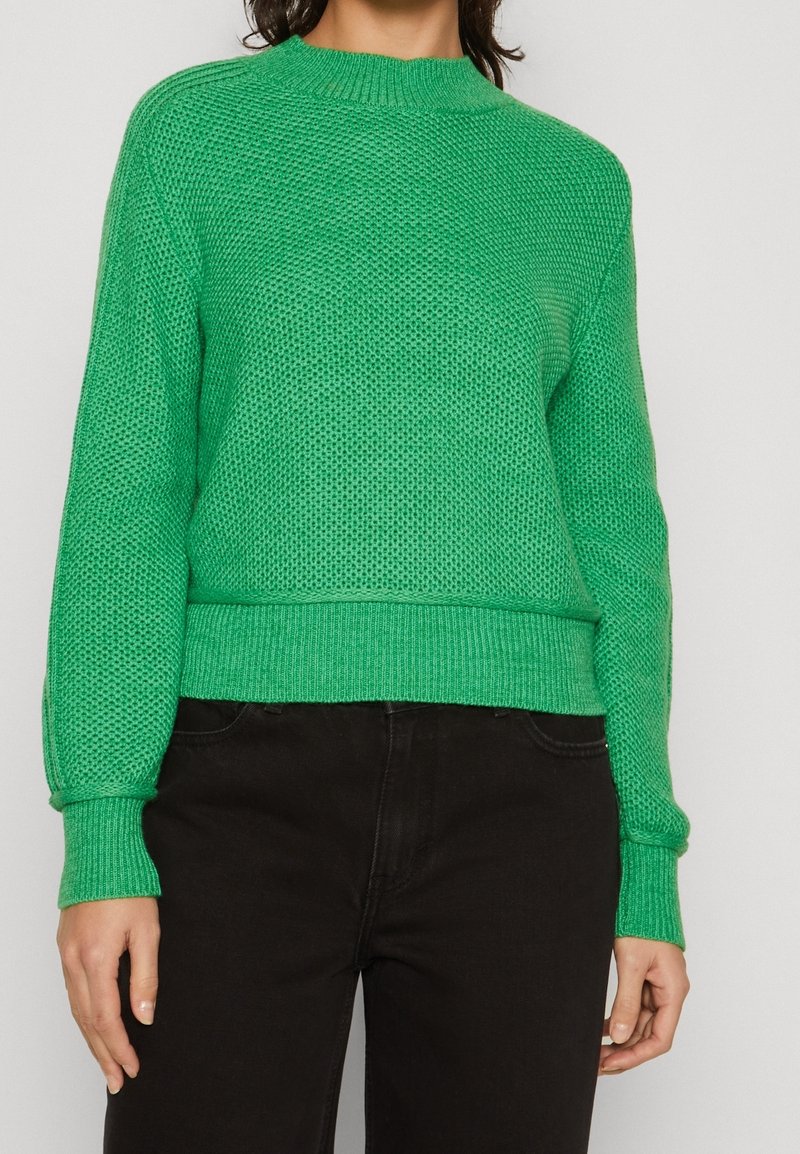 Femme portant un pull en maille texturé vert vif avec poignets et ourlet côtelés, associé à un pantalon noir sur un fond uni.