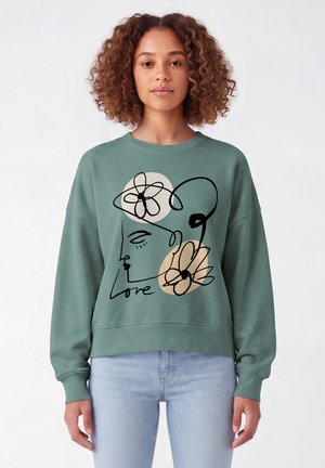 Frau mit lockigem Haar, die einen grünen Pullover mit abstraktem Gesicht und Blumenlinienkunst mit dem Wort "Love," trägt, kombiniert mit hellblauen Jeans.