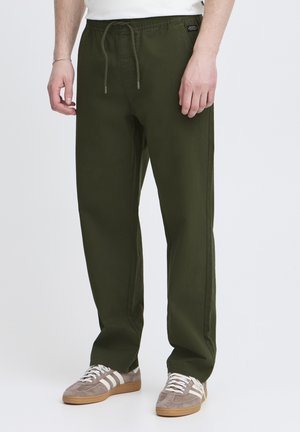Green cotton blend pants with an elastic waistband, adjustable drawstring, side pockets, and a relaxed fit. Worn with gray sneakers.
