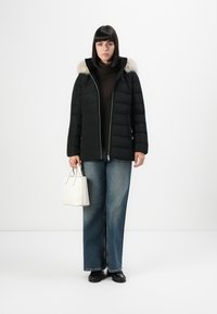 Donna che indossa un giubbotto piumino nero con cappuccio in pelliccia, maglione dolcevita scuro, jeans larghi, scarpe nere, mentre tiene una piccola borsa bianca, in piedi su uno sfondo bianco.