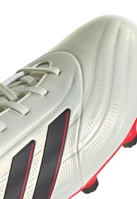 Botín de fútbol gris claro con acentos en negro y rojo, que presenta un material sintético texturizado, cordones y un diseño texturizado y aerodinámico.