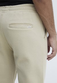 Beige Sweatpants mit einer Gesäßtasche, die ein genähtes Etikett aufweist. Sie bestehen aus weichem Stoff, haben einen elastischen Bund und einen geraden Schnitt.
