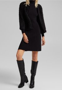 Femme portant une veste noire texturée sur une robe noire côtelée à col roulé, assortie à des bottes en cuir noires montant jusqu'aux genoux, debout devant un fond gris.