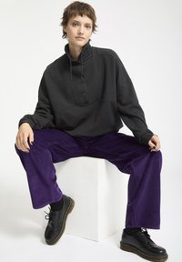 Felpa nera oversize con colletto alto e bottoni, abbinata a pantaloni in corduroy viola a coste e scarpe nere con lacci.