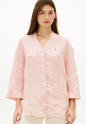 Mujer con cabello largo y castaño lleva una camisa de rayas verticales rosas y blancas con botones y mangas tres cuartos, y pantalones de color claro.