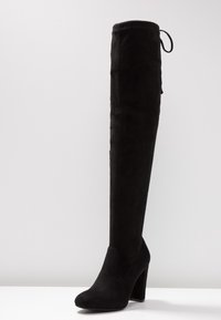 Černé kozačky do výšky kolen vyrobené z měkké semišové kůže s hladkým povrchem, kulatou špičkou a vysokým blokovým podpatkem. Obsahují nastavitelné šněrování na vrchu.