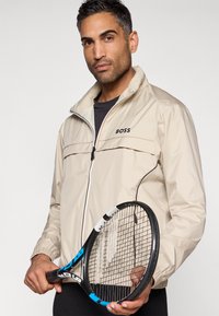Bēšs viegls džemperis ar rāvējslēdzēja aizdari, BOSS logo uz krūtīm un elastīgiem aprocēm. Modelis tur melnu un zilu tenisa raketi.