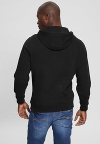 Black cotton hoodie with a rear view, featuring a large, soft hood and ribbed cuffs, paired with blue denim jeans. Simple, clean design.