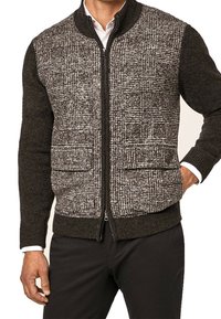 Uomo che indossa un cardigan con zip, con pannelli frontali a scacchi bianco e nero e maniche nere solide, abbinato a pantaloni neri e una camicia bianca.