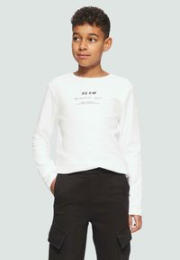 Jongen in een wit longsleeve shirt met tekst en zwarte cargobroek, staand met één hand in zijn zak, kijkend opzij.