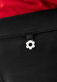 Juodas audinio juosmuo su sidabrinės spalvos gėlės formos žyma, kurioje yra centrinė anga, kontrastuojanti su raudona viršaus dalimi viršuje.