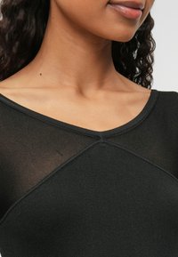 Czarny dopasowany top z prążkowanego materiału z dekoltem w kształcie litery V; posiada przezroczyste panele i trójkątny detal szwu na ramieniu.