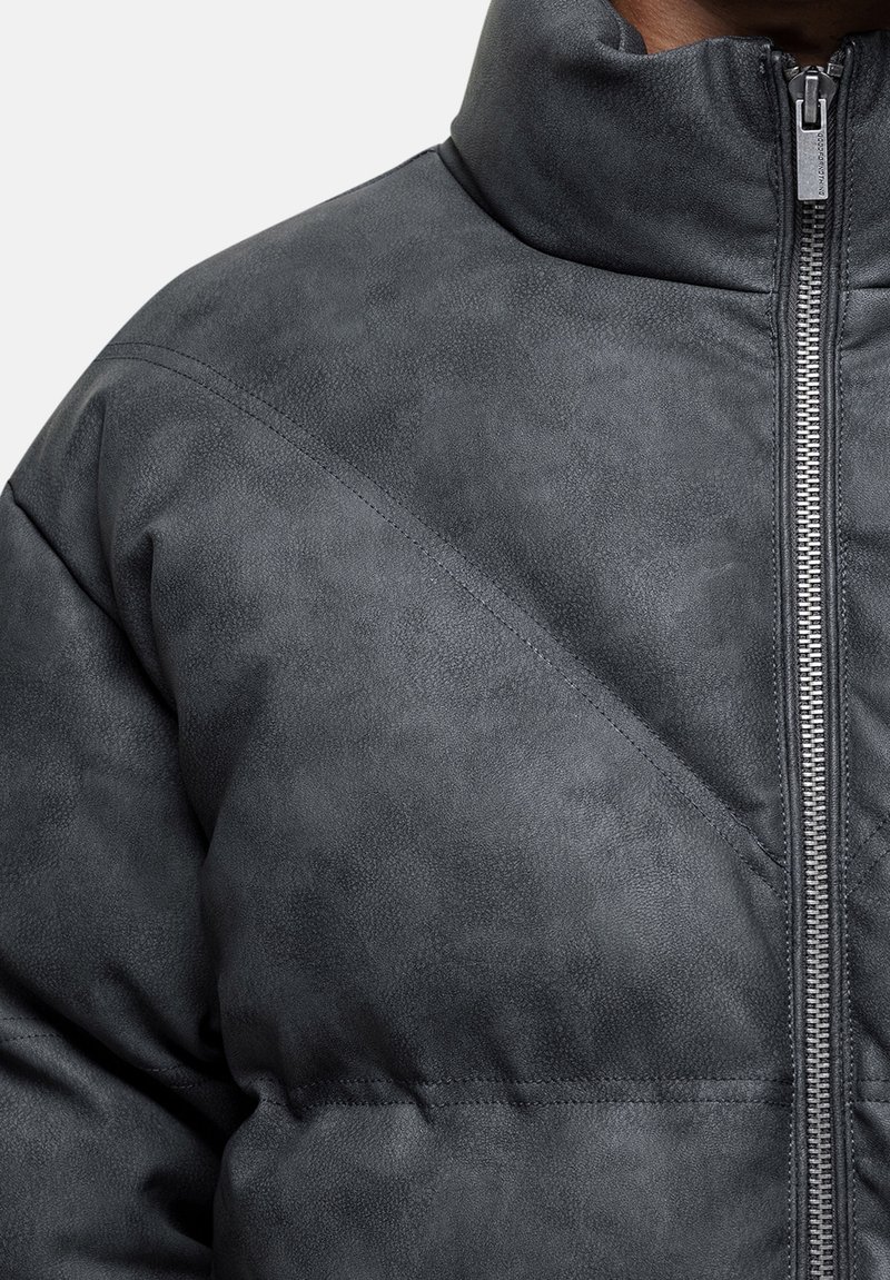 Graue Kunstlederjacke mit hohem Kragen, diagonalen Nähdetails und einem Reißverschluss vorne. Die Textur wirkt glatt mit einem leichten Glanz.