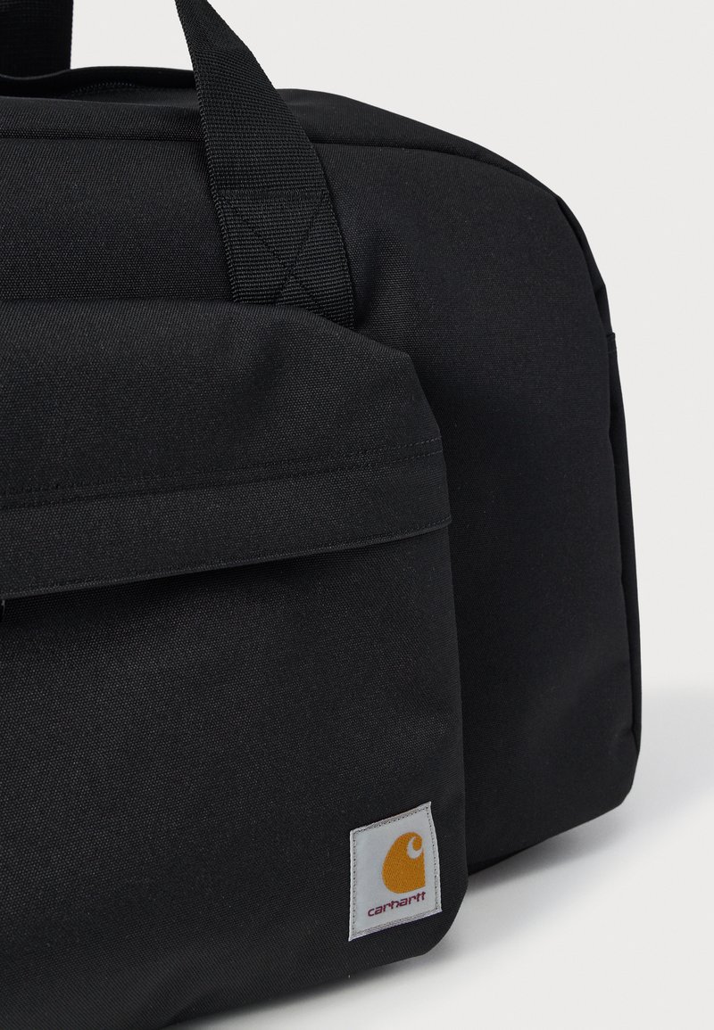 Close-up van een zwarte Carhartt-tas met een ritsvak aan de voorkant en een stoffen handvat tegen een effen lichte achtergrond.