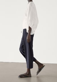 Blusa bianca, morbida e ampia con maniche a sbuffo, abbinata a pantaloni cropped blu scuro e infradito nere, su uno sfondo neutro.