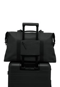 Sac de sport noir avec une texture, doté de deux poignées et d'une poche zippée, posé sur une valise à roulettes noire.