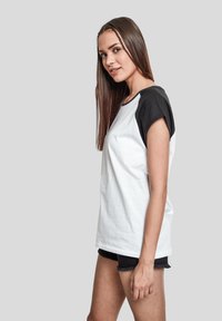 Camiseta blanca de algodón con mangas raglán negras, escote redondo y mangas cortas. Llevada con pantalones cortos de mezclilla negros.