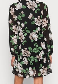 Musta kukkakuvioinen mekko, jossa on pitkät hihat, kerroksellinen hame ja kermanväristen kukkien sekä vihreiden lehtien kuvio läpinäkyvällä kankaalla.
