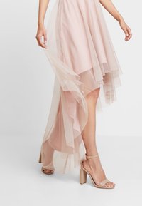 Jasnoróżowa sukienka z warstwowym tiulem, z asymetrycznym dołem. Modelka ma na sobie cieliste sandały na obcasie. Teksturowany materiał dodaje głębi.