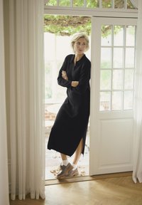 Black long-sleeve dress with a side slit, worn with gray lace-up shoes and white socks, standing by a partially open door with glass panes.