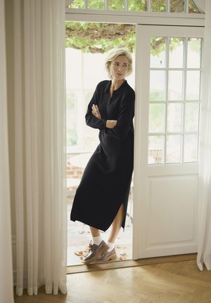 Schwarzes Langarmkleid mit einem seitlichen Schlitz, getragen mit grauen Schnürschuhen und weißen Socken, steht an einer teilweise geöffneten Tür mit Glasfenstern.