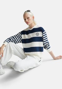 Felpa corta a righe blu navy e bianche con logo centrale, abbinata a pantaloni della tuta bianchi e scarpe bianche chunky. Tessuto dalla texture liscia.