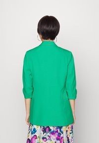 Zelené strukturované sako s tříčtvrtečními rukávy, které má klopový límec a boční kapsy, v kombinaci s květinovým vzorem sukně.