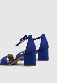 Sandalias de tacón de ante azul con talón cerrado, tira en el tobillo y diseño de punta abierta. Tacón grueso y suela lisa.