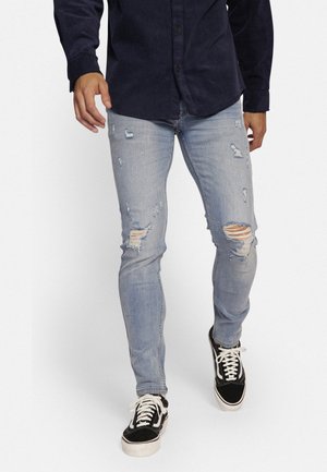 Lichtblauwe distressed skinny jeans met scheuren op de knieën, gecombineerd met zwart-witte sneakers en een marineblauwe overhemden.