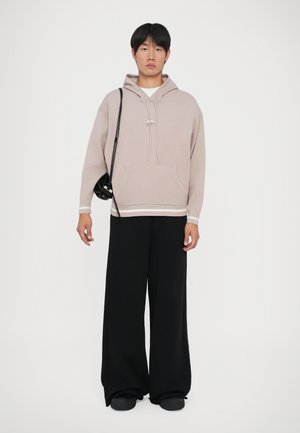 Joven de pie, vistiendo una sudadera beige, pantalones anchos negros, zapatos negros y llevando una pequeña bolsa negra de hombro, contra un fondo blanco.