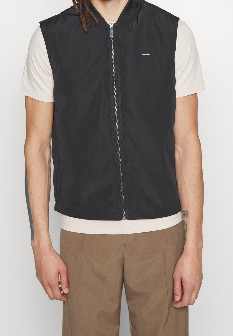 Personne portant une veste noire sans manches à fermeture éclair sur une chemise beige à manches courtes et un pantalon marron, debout avec les bras détendus le long du corps.