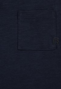 Tumma laivastonsininen kangas, jossa on neliönmuotoinen rintatasku ja sen alakulmassa oikealla pieni musta kirjailtu "R".