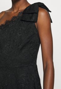 Svart spetsklänning med design på ena axeln, med ett strukturerat blommönster och en rosettdetalj på axeln.